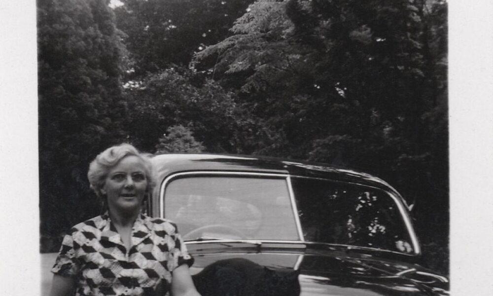 Vrouw in geruite jurk naast een klassieke auto, zwart-witfoto