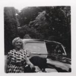 Vrouw in geruite jurk naast een klassieke auto, zwart-witfoto